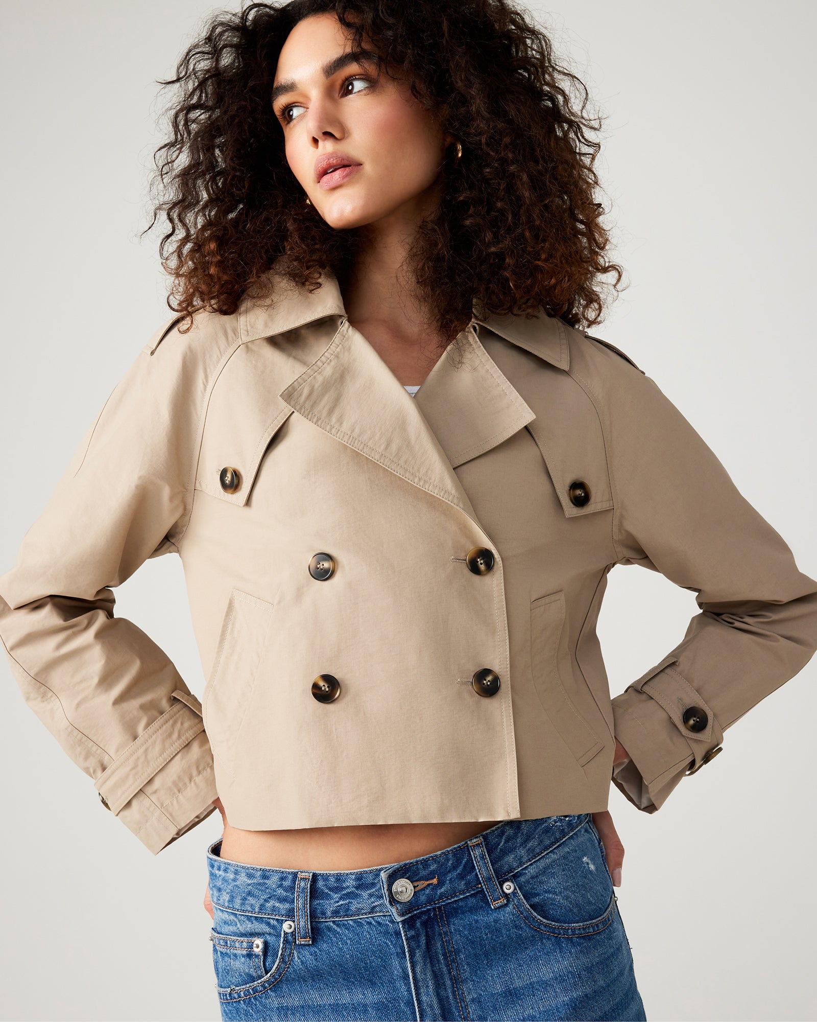 Front view of khaki cropped trench jacket on model in an urban setting.